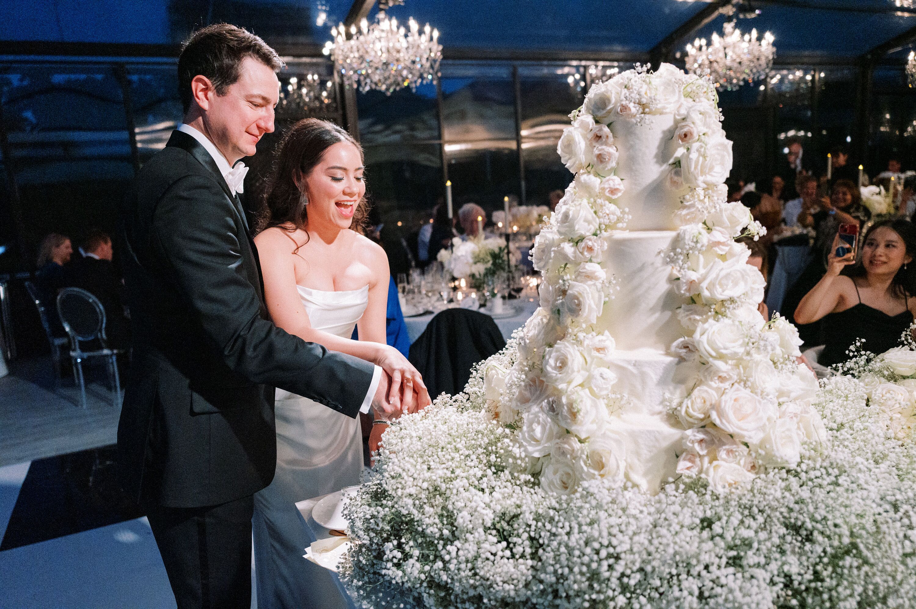 Couple Cutting Luxurious Multi-Tiered Wedding Cake With Fresh White Flowers