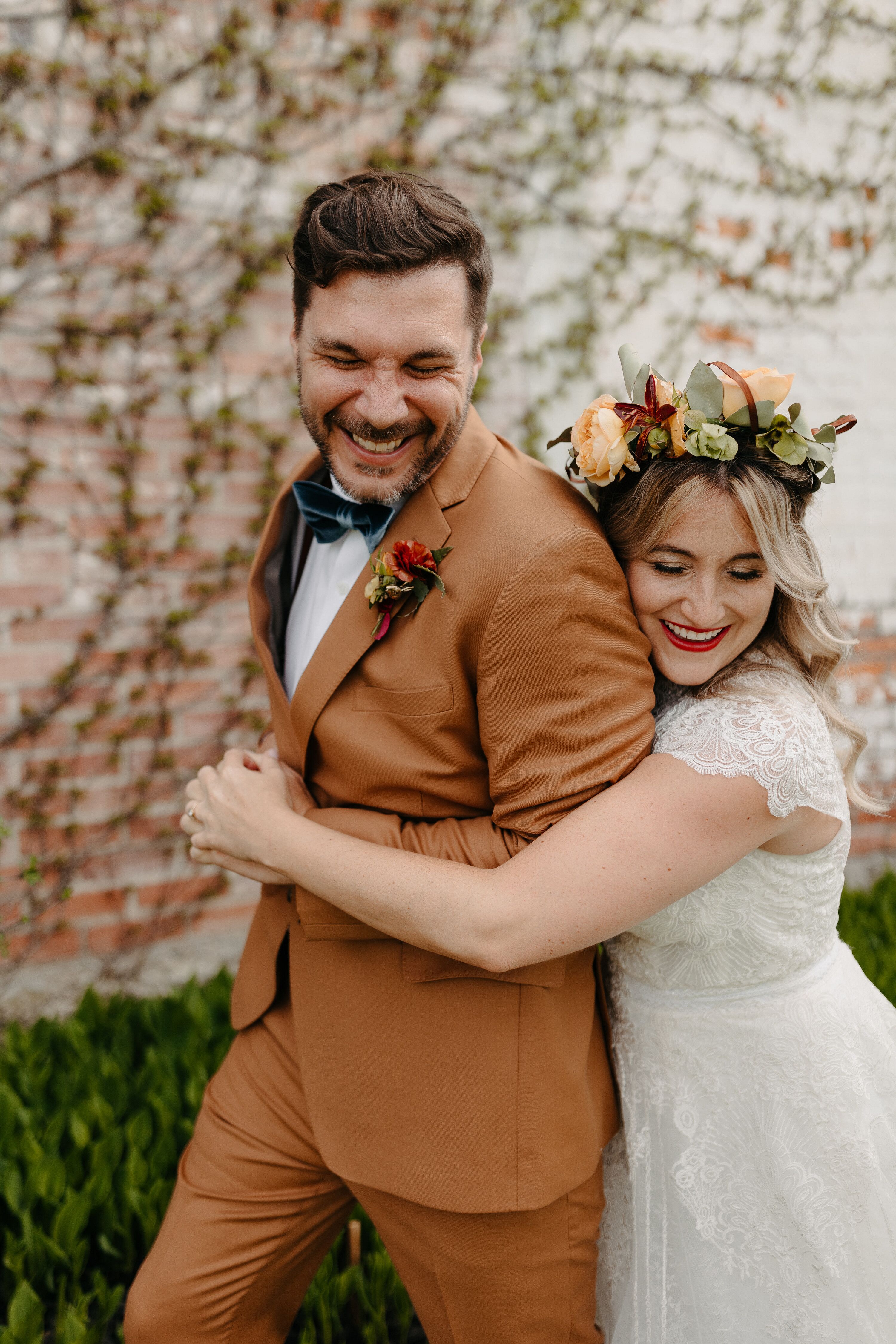 Bride in Lace Dress and Flower Crown Hugs Groom in Burnt Orange Suit ...